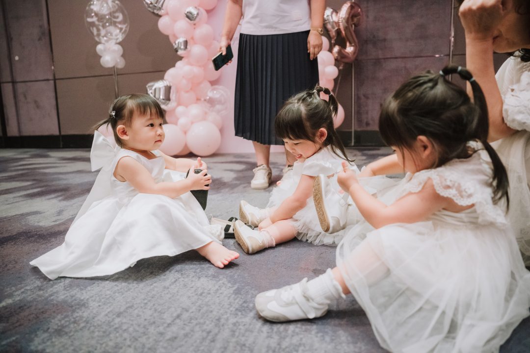 三個穿白洋裝的小女孩坐在地毯上脫鞋子互相玩耍。