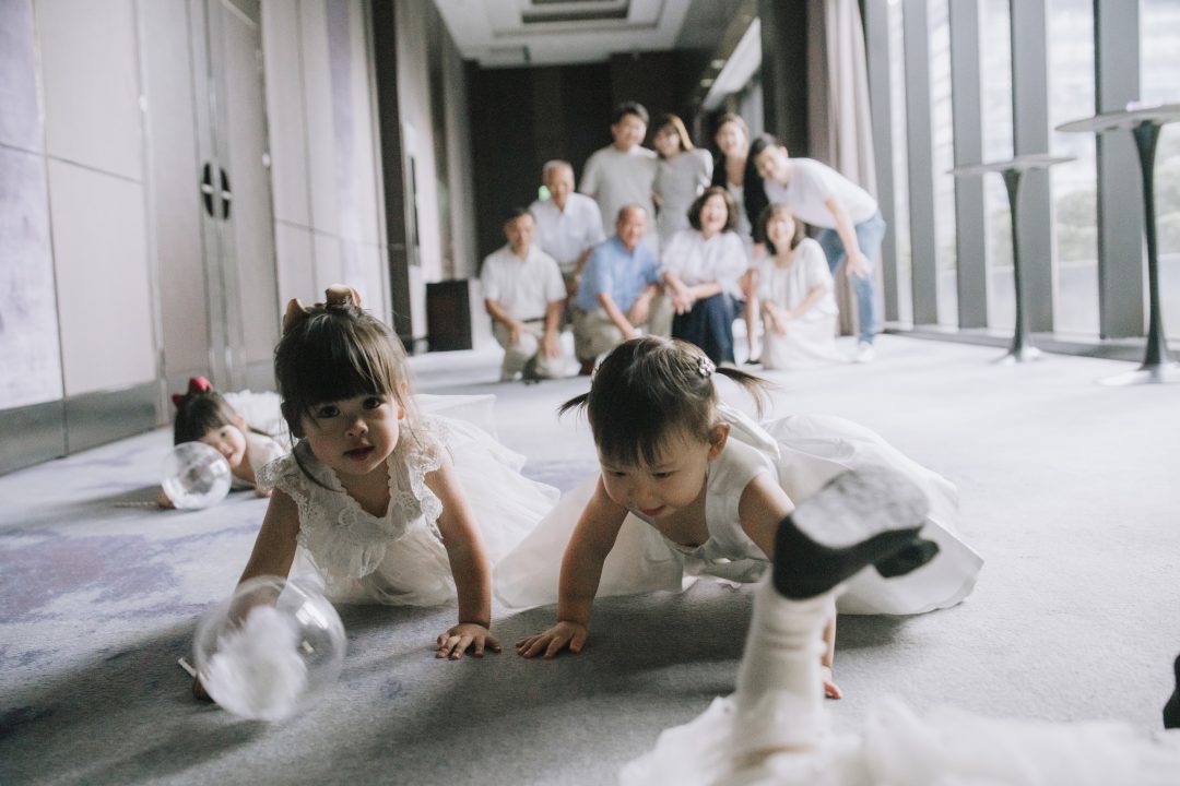 雙胞胎姊妹趴在灰色地毯上向前爬，遠處親友溫柔注視