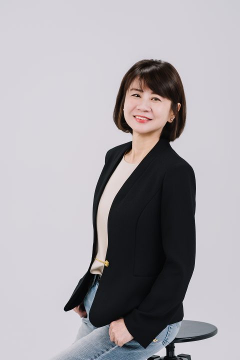 素灰背景前穿黑西裝與牛仔褲，坐在高腳椅上側身微笑