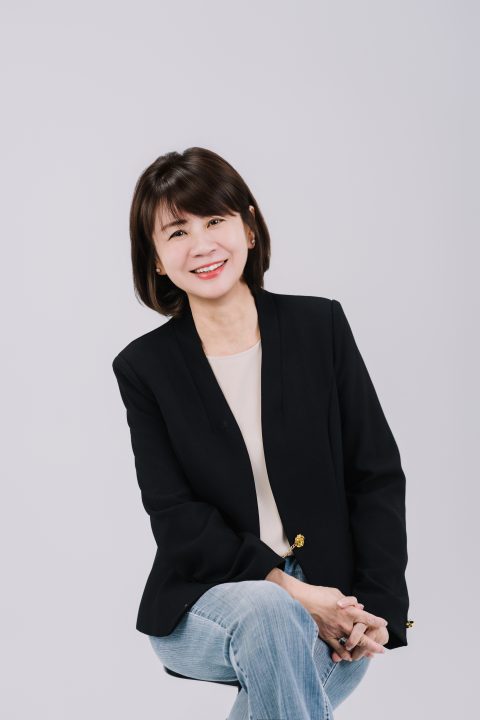 上身微微前傾且雙手交疊在膝上，穿著黑色西裝開懷露齒大笑