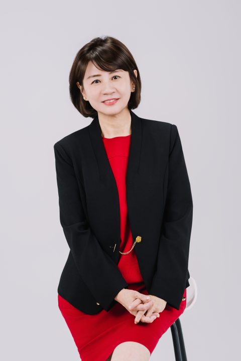黑西裝內搭紅色裙裝，雙手十指交扣端坐在純灰背景高腳椅上