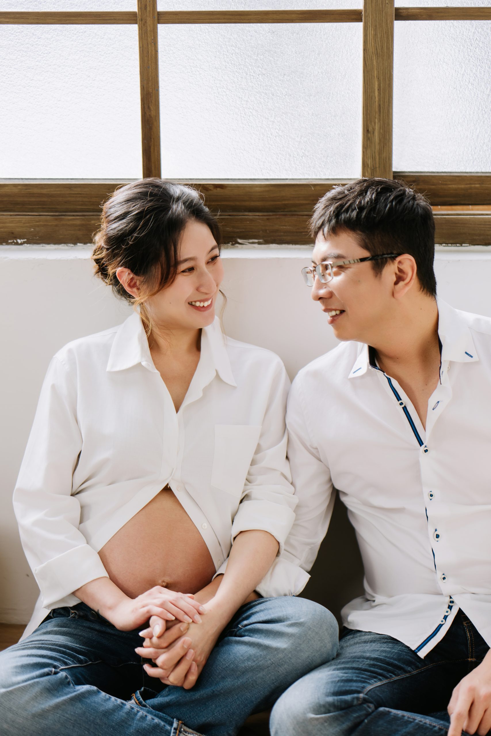 席地而坐的夫妻雙手交握相視而笑，白色襯衫下露出圓潤的孕肚。