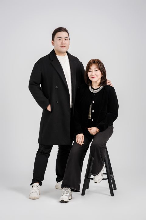穿著黑色毛呢長大衣的兒子站在椅旁，一手搭在盛裝母親的肩上