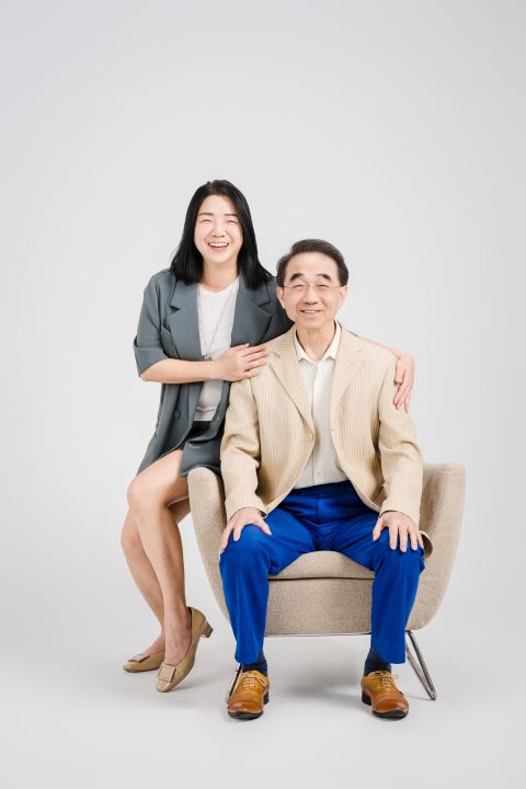 女兒側靠在單人布沙發扶手旁開懷大笑，手輕搭在父親肩上合照