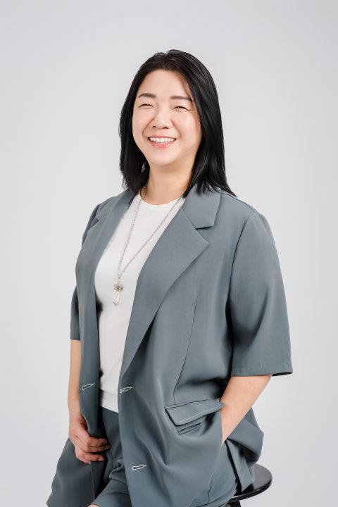 身穿灰藍色西裝套裝的女性坐在圓凳上，雙手插口袋露出自信笑容