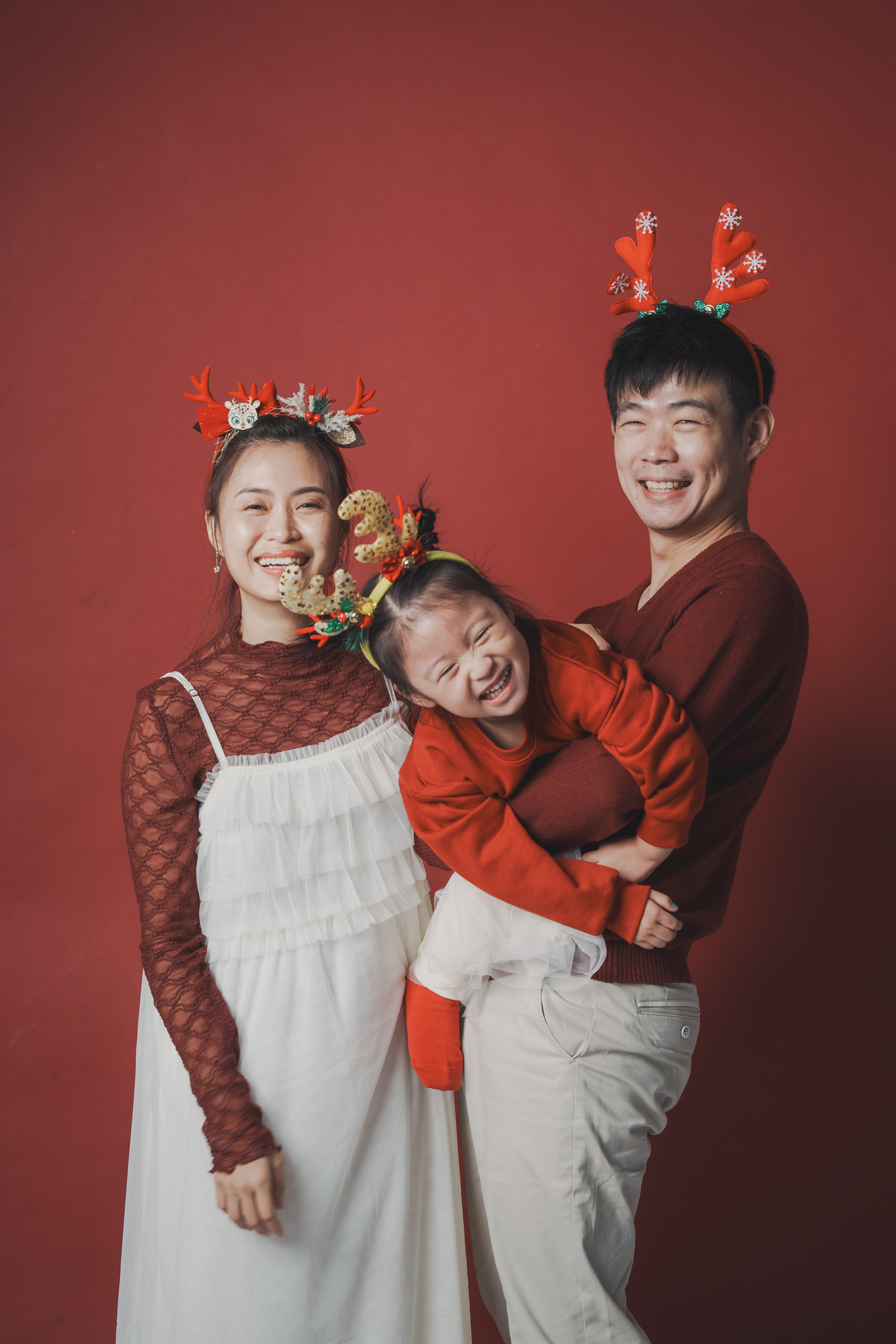 紅色背景前爸爸抱起大笑的女兒，全家戴著聖誕髮飾合影